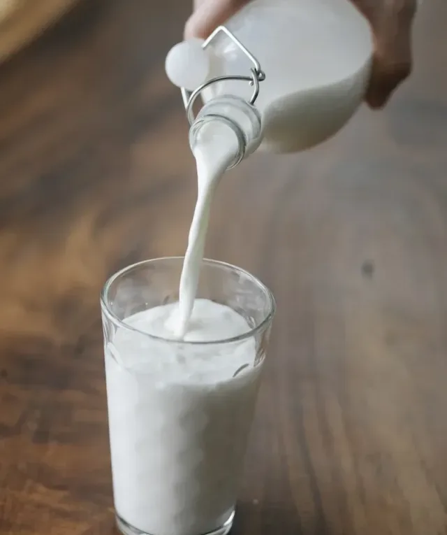 Crop anonymous person with milk bottle pouring milk into ornamental transparent glass on wooden table at home